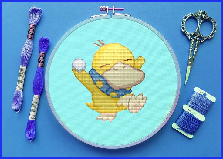 a cross stitch kit with scissors, thread and needle on a blue surface next to it is a yellow teddy bear holding a ball of yarn