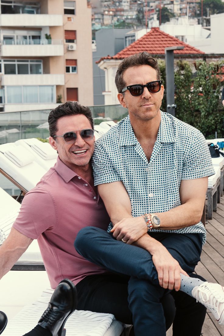 two men sitting next to each other on top of a wooden bench in front of buildings