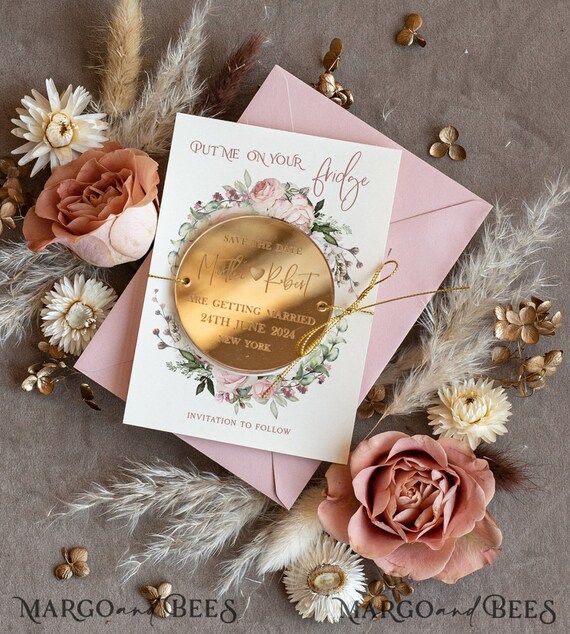 two pink and gold wedding cards on top of some feathers with flowers in the background