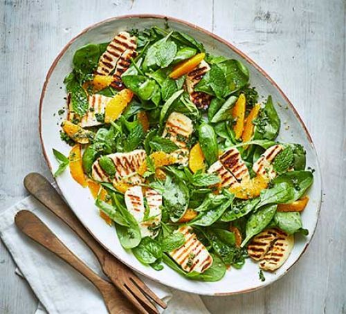 a salad with grilled chicken and oranges on it in a white bowl next to two wooden utensils