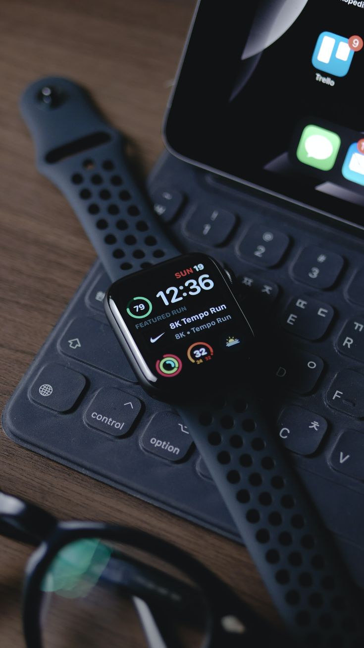 an apple watch sitting on top of a keyboard next to a smart phone and eyeglasses