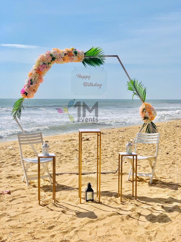 two white chairs sitting on top of a sandy beach next to an arch decorated with flowers