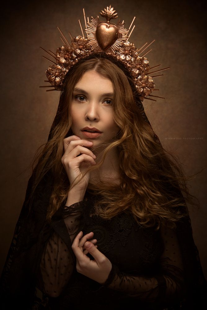 a woman with long hair wearing a crown and holding her hand to her chin, in front of a dark background