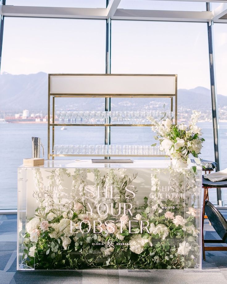 a clear box with flowers and greenery on the front sits in front of a large window