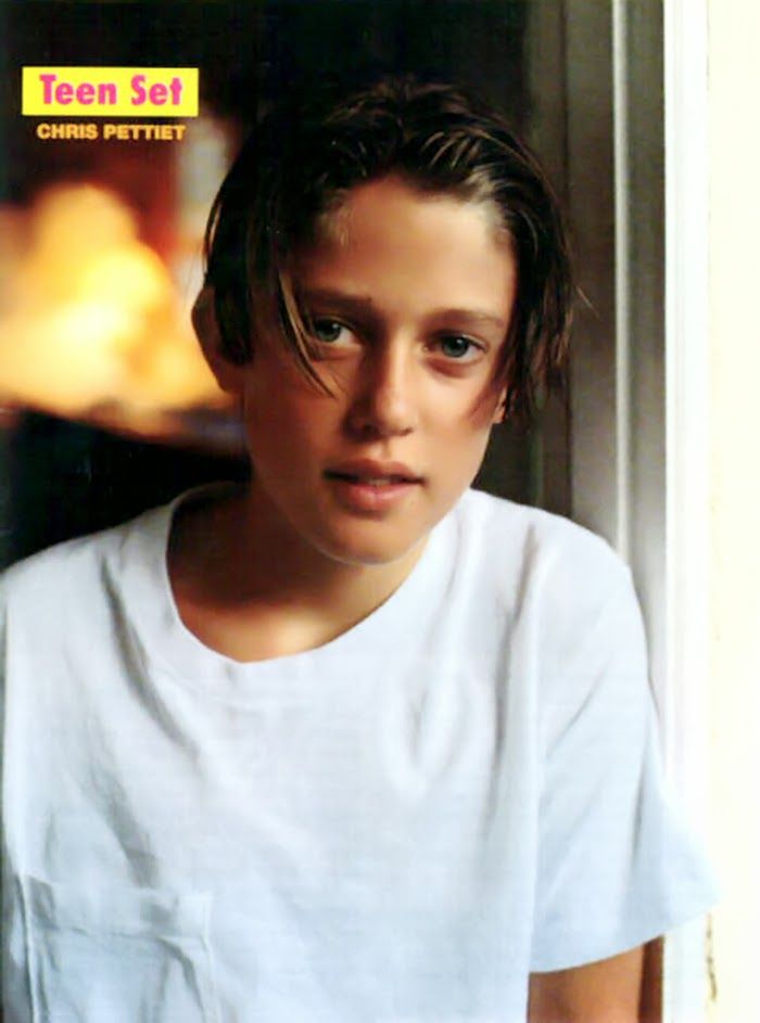 a young boy is looking at the camera while wearing a white t - shirt and standing in front of a window