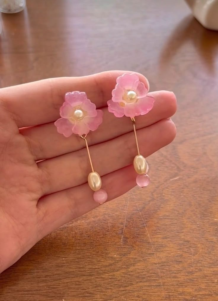 a person is holding some pink flowers and pearls on their earring wires, while they are sitting on a wooden table