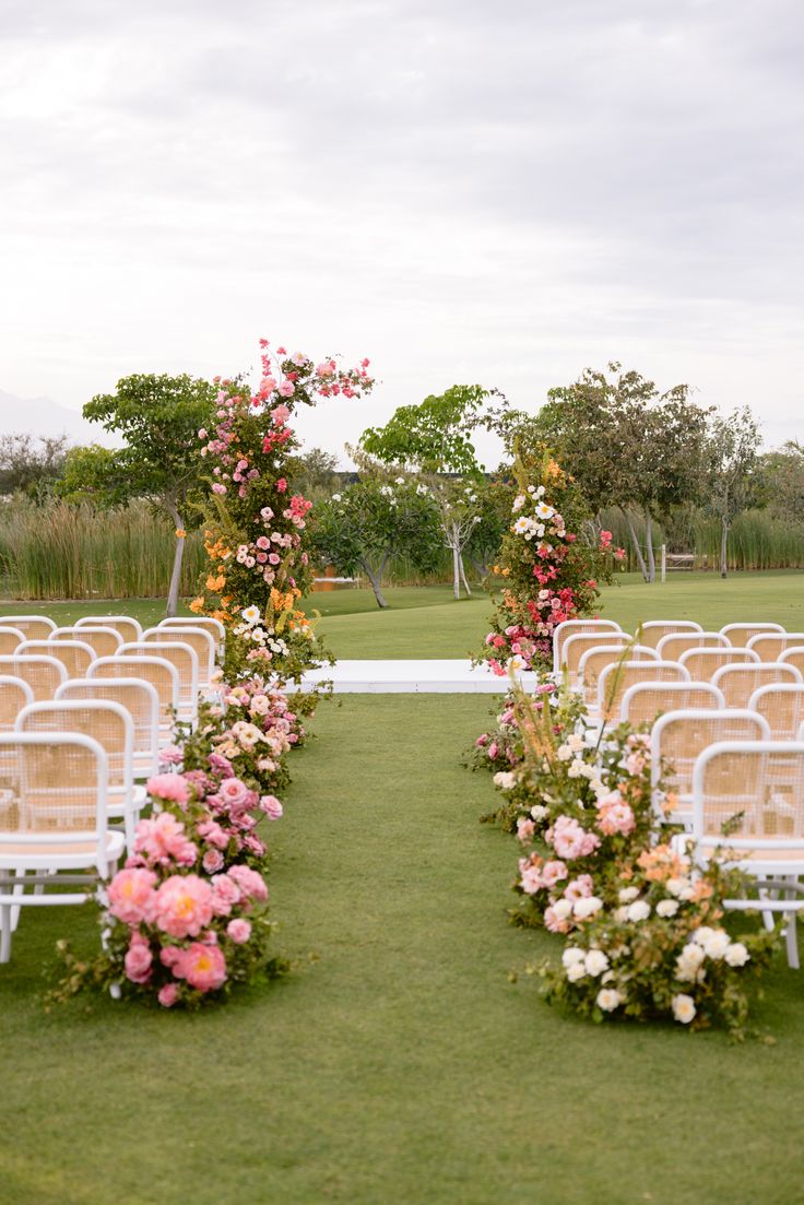 an outdoor ceremony set up with white chairs and pink flowers on the aisle, surrounded by greenery