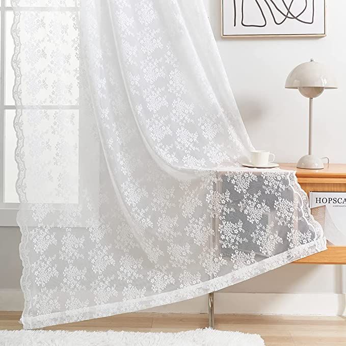 a white lace curtain hanging in front of a window next to a table with a lamp on it