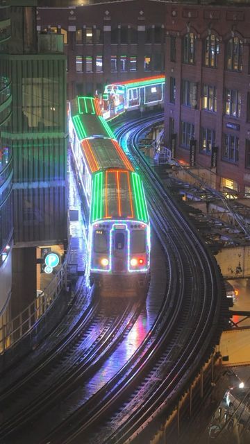 a train traveling down tracks next to tall buildings in a city at night with lights on