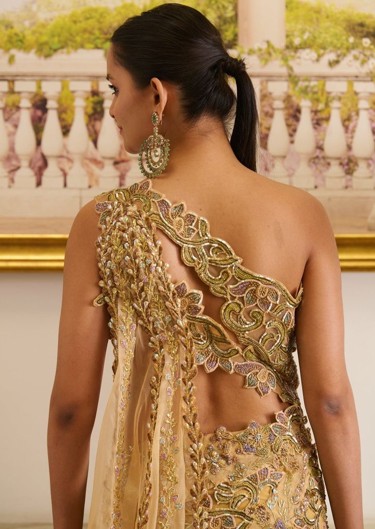 Golden hour is a stunning gold and mustard pre-stitched saree, which features a halter-neck blouse, floral jacket, and a pre-draped skirt. The cut-work adorning the blouse, with olive green floral embroidery, presents sensual glamour, paired up with a pre-draped skirt featuring a modest slit and embellished with floral motifs accentuated with sequins and crystal detailing. Halter-neck blouse adorned with floral embroidery and cut-work detailing as well. Floral jacket accentuated with sparkling beads, crystals, and sequins. Pre-stitched skirt featuring a modest slit embellished with defining the modern, sensual romance of shimmery soirées, this pre-draped rendition of a saree speaks chic glam on point. The structured silhouette synchronizes with the flower motifs embellished with zari and z Desi Ootd, Gown Saree, Mustard Outfits, Skirt Saree, Halter Neck Blouses, Stitched Saree, Shoulder Dresses, Flower Motifs, Draped Skirt