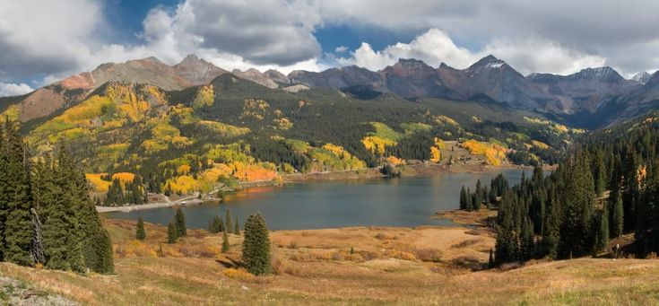 a lake surrounded by mountains and trees with fall colors in the foreground on a cloudy day