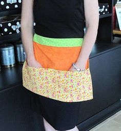 a woman standing in front of a counter holding an orange and green apron