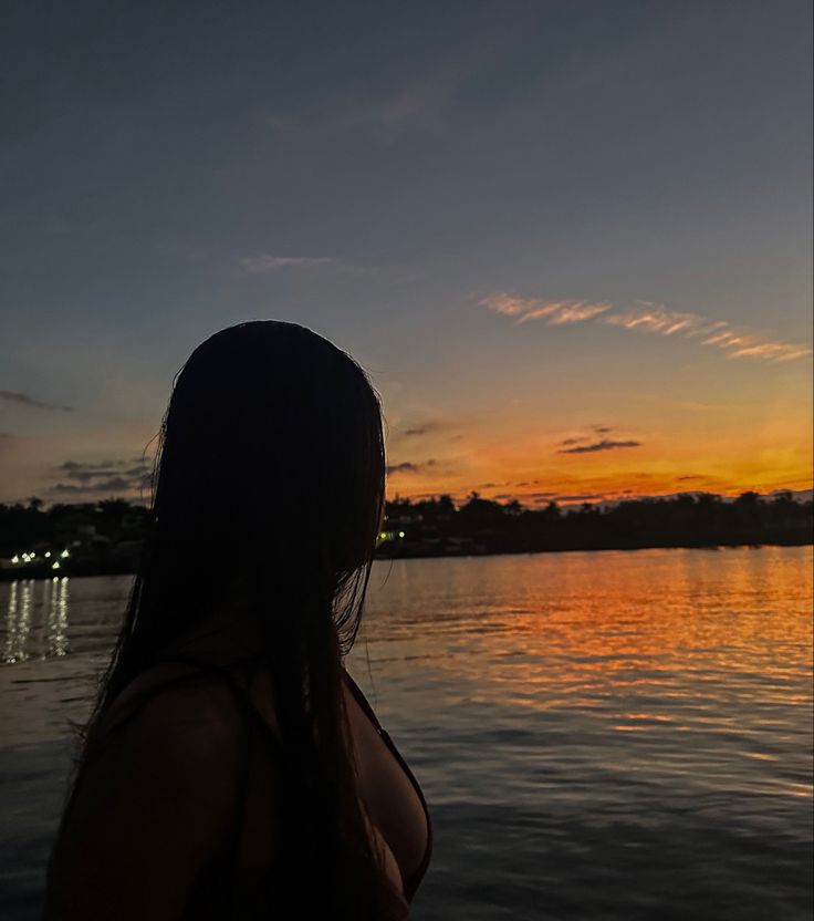 a woman standing on the edge of a body of water at sunset with her back to the camera