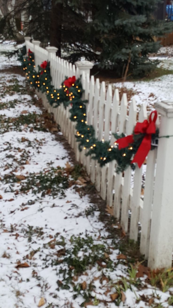 a white picket fence decorated with christmas lights and wreaths in the snow next to a pine tree