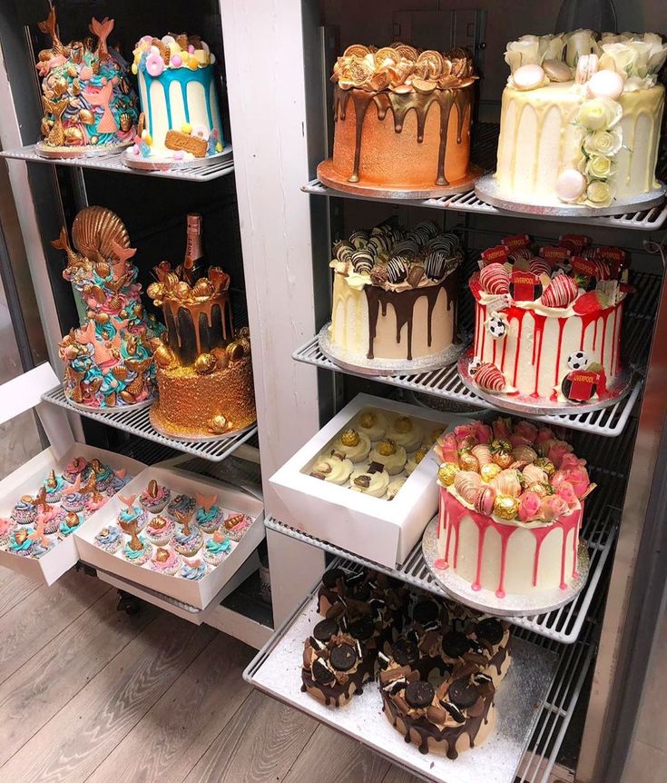 many different cakes and pastries are on shelves in a bakery display case, all decorated with colorful icing