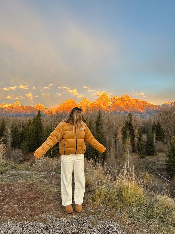 a woman standing on top of a grass covered field next to mountains with trees in the background