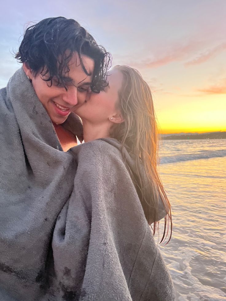 a man and woman wrapped in a blanket on the beach at sunset, one is kissing the other's forehead