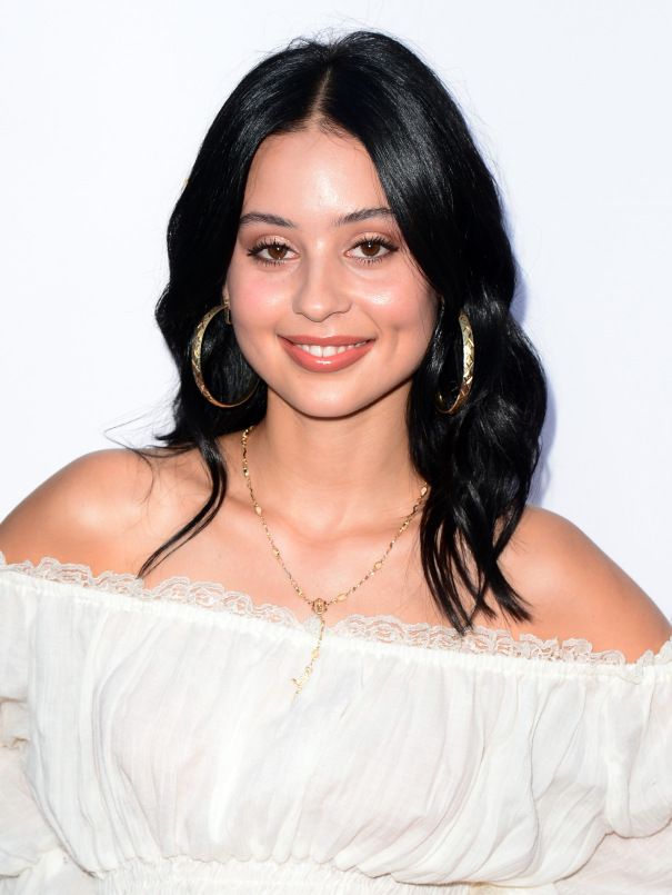 a woman with long black hair wearing large hoop earrings and a white dress smiling at the camera