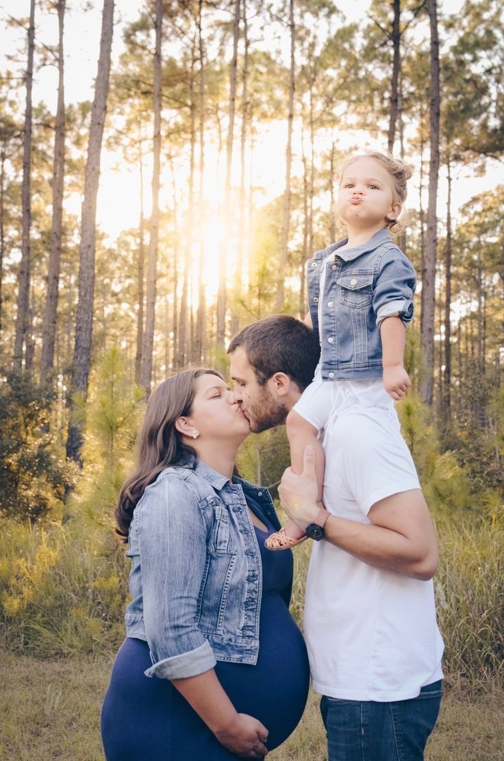 a pregnant woman kissing her husband's belly as they stand in front of trees