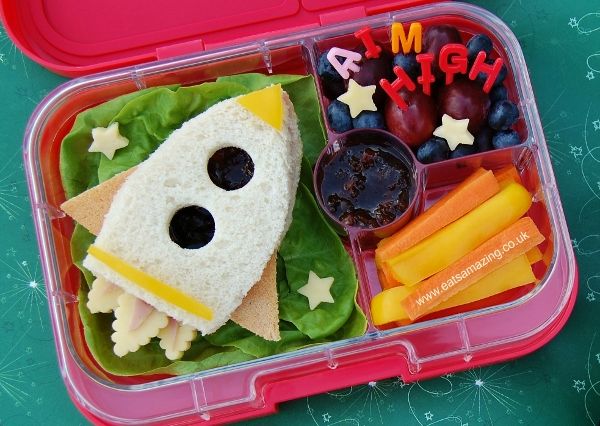 a lunch box with a sandwich, fruit and veggies in the shape of a rocket ship