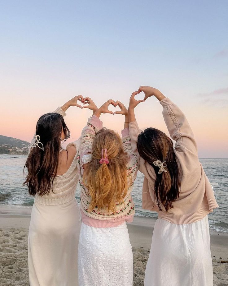 three women standing on top of a beach holding their hands in the shape of a heart