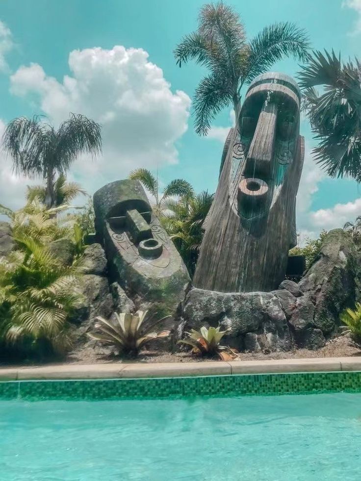 a large wooden sculpture sitting next to a pool in a lush green forest filled with palm trees