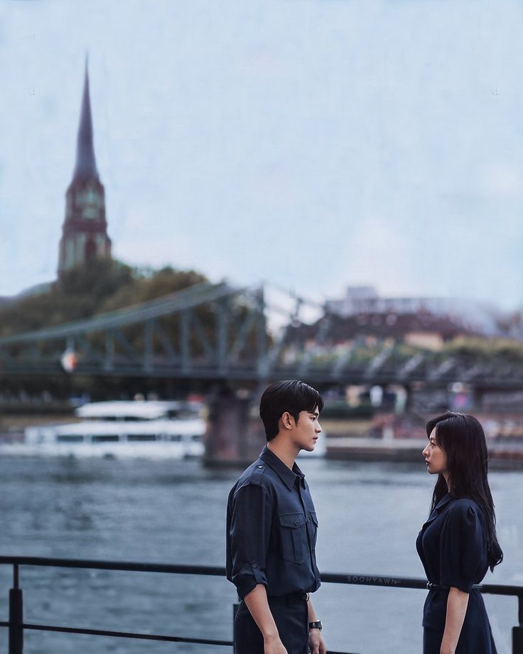 a man and woman standing next to each other on a bridge over water with a city in the background