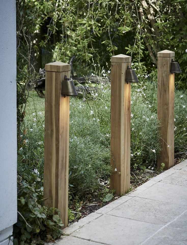 three wooden posts with lights on them in front of some bushes and trees, along side a sidewalk