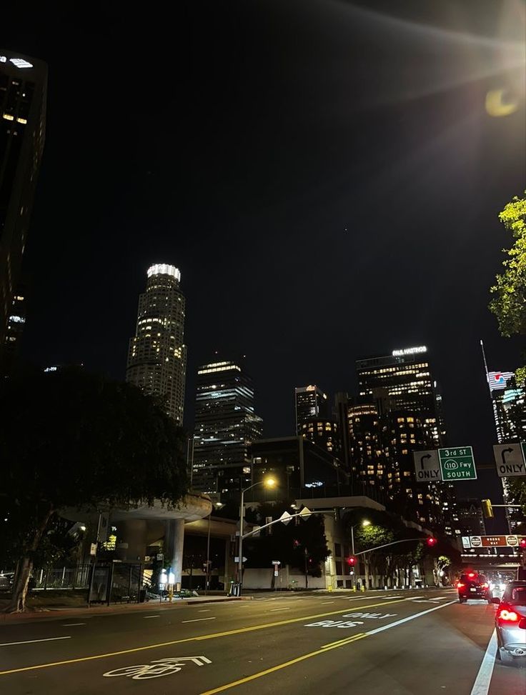 a city street at night with cars driving on the road and skyscrapers in the background