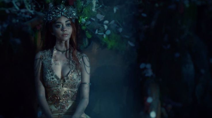 a woman with red hair wearing a gold dress and a crown standing in the woods