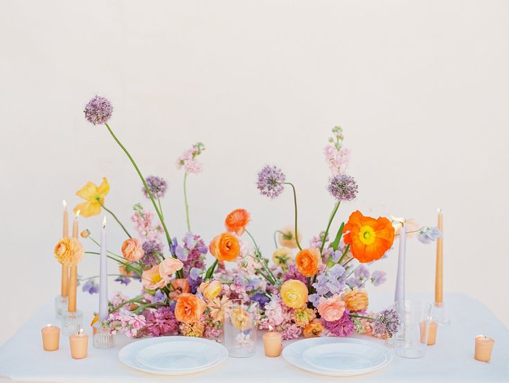 an arrangement of colorful flowers and candles on a white table cloth with blue place settings