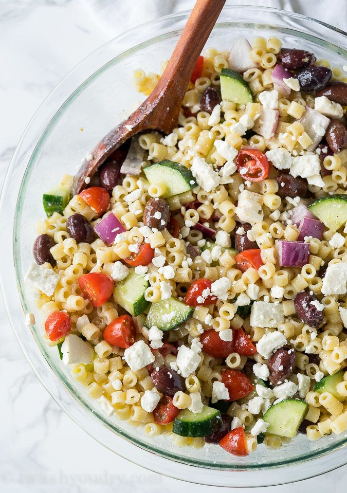 a glass bowl filled with pasta salad on top of a white marble countertop next to a wooden spoon