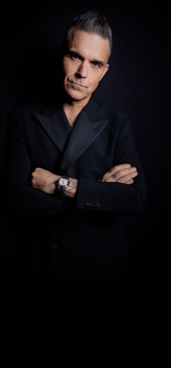 a man in a black suit with his arms crossed and looking at the camera while wearing a watch