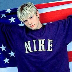 a young man with blonde hair standing in front of an american flag and holding his arms behind his head