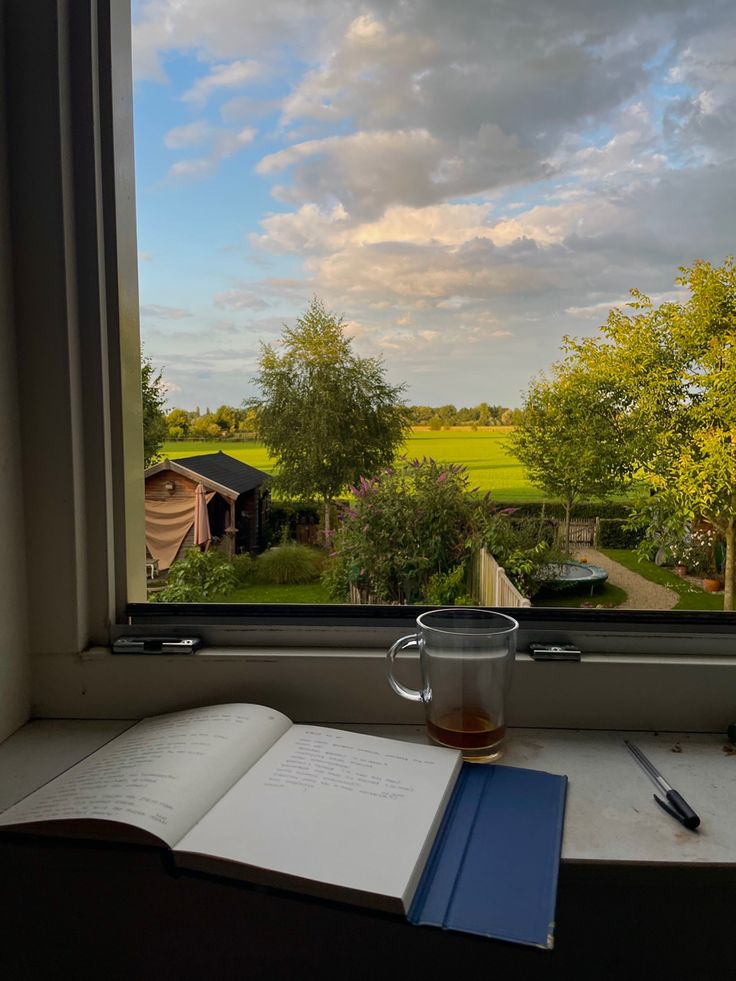 an open book and cup on a window sill with a view of the countryside