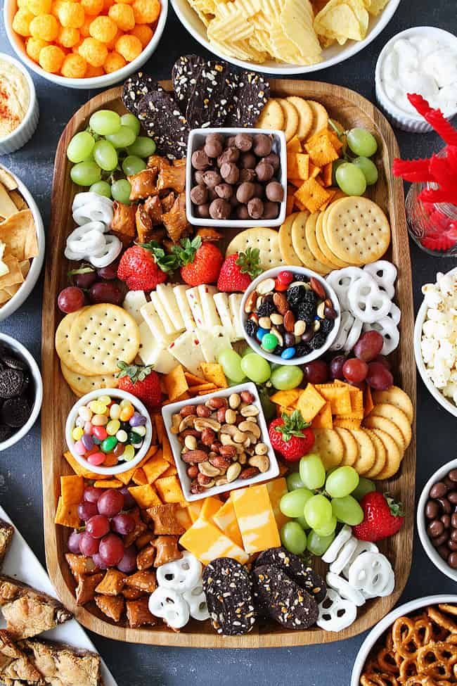 a platter filled with cheese, crackers, fruit and nuts is surrounded by other snacks