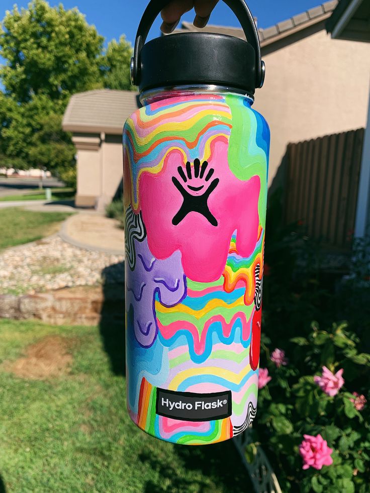 a colorful water bottle hanging from a metal hook in front of a house with pink flowers