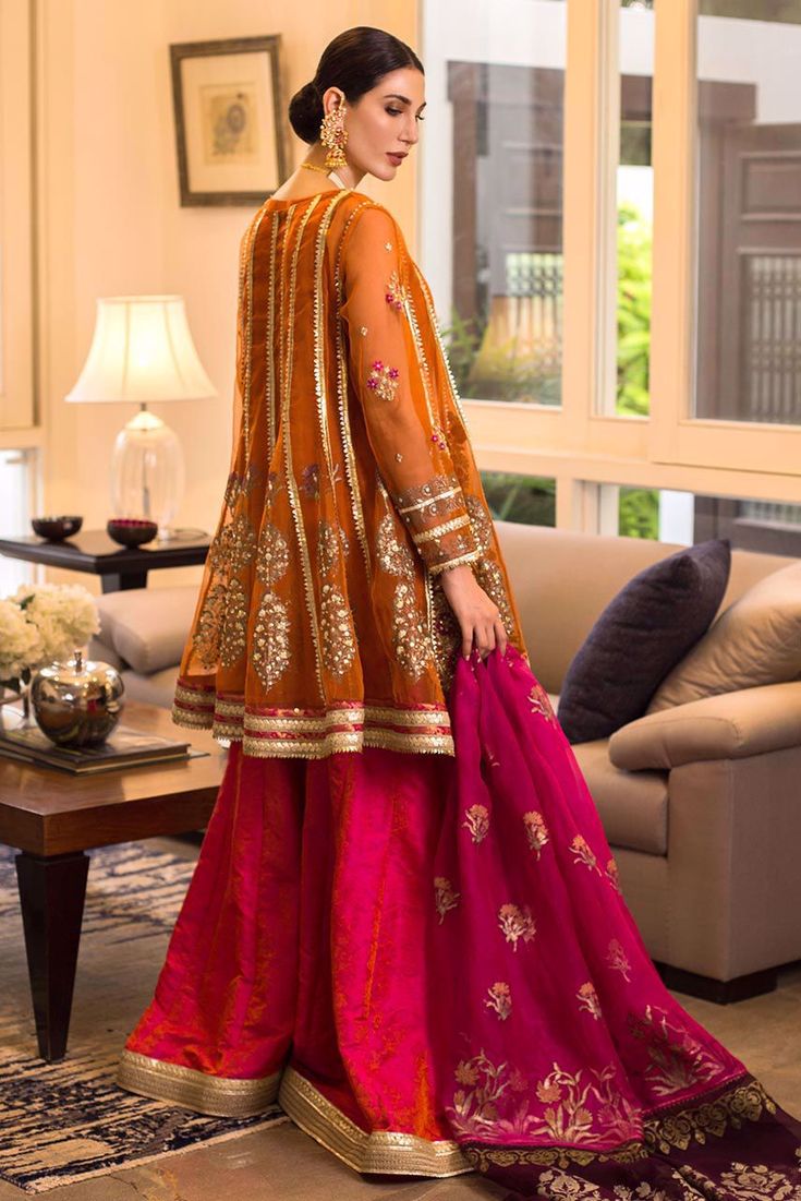 a woman in an orange and pink outfit stands near a couch, looking off to the side