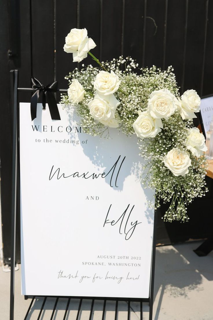 Classy welcome sign adorned by upper right corner baby's breath and white rose design. White Roses Baby’s Breath Centerpiece, Wedding Signs Flowers, Baby’s Breath And White Rose Centerpieces, Wedding Easel Flowers, White Wedding Ideas Decoration, Wedding Sign Flower Arrangement, Baby Breathe Weddings, White Rose And Baby’s Breath, White Flower Wedding Ceremony