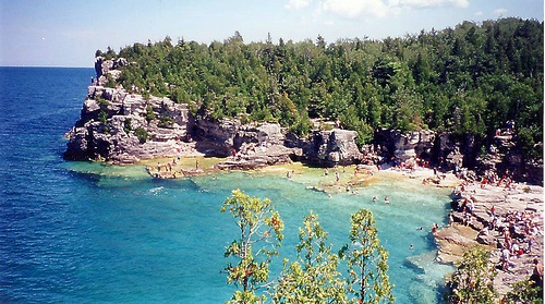 people are swimming in the blue water next to some rocks and trees on the shore