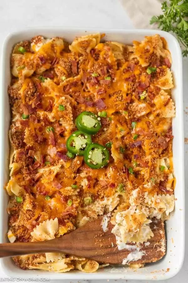 a casserole dish with cheese, bacon and green peppers in it next to a wooden spoon