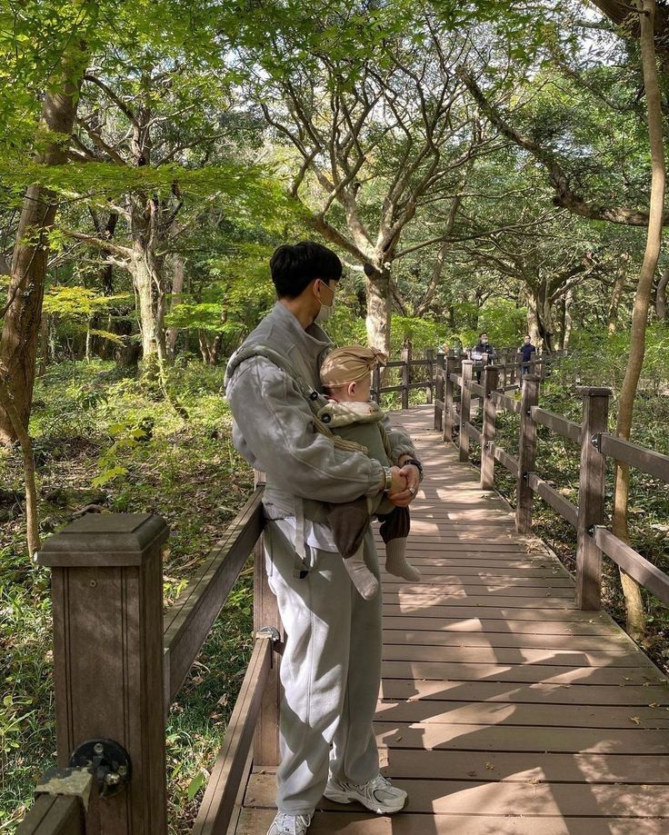 a man holding a baby while standing on a wooden walkway in the woods with trees