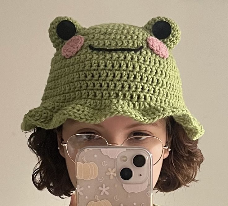 a woman taking a selfie with her phone wearing a crocheted frog hat