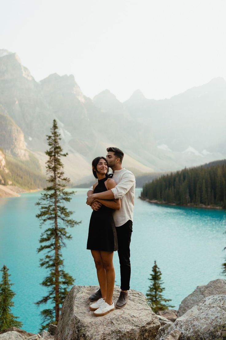 a man and woman standing on top of a rock next to a lake