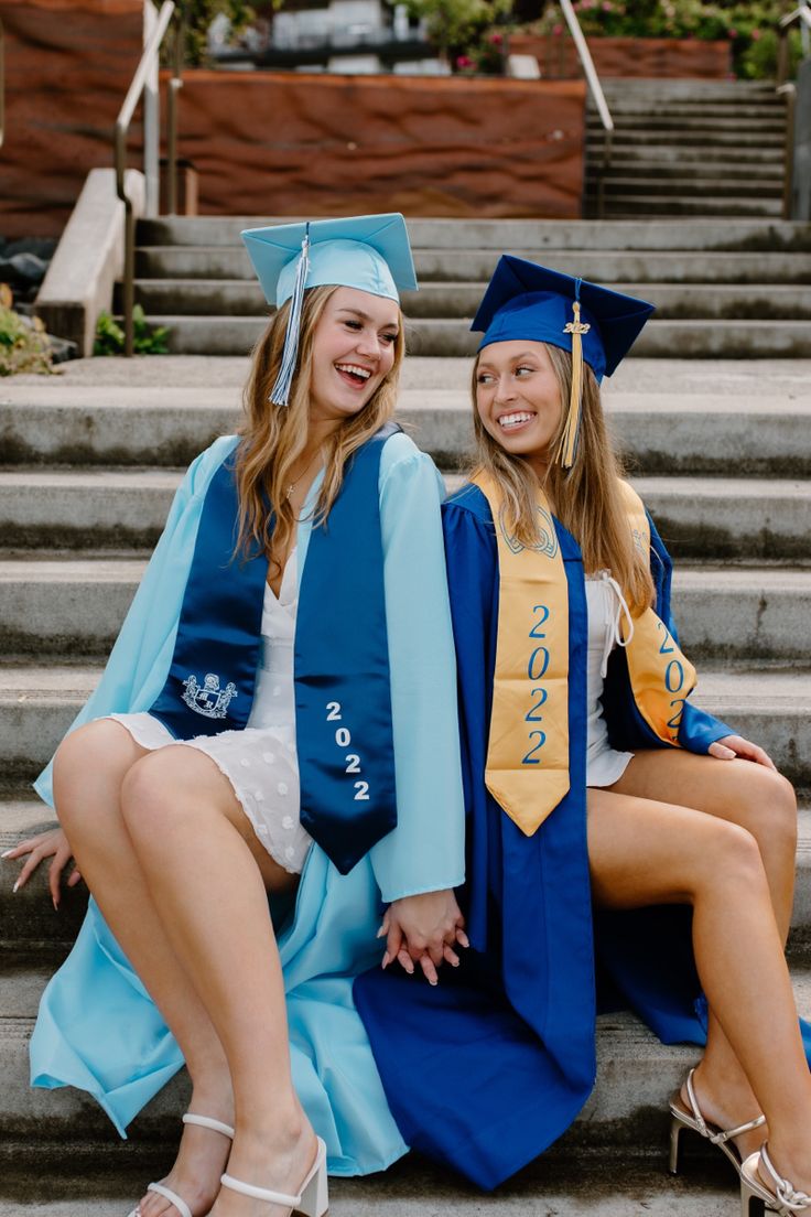 two young women in graduation gowns sitting on the steps with their arms around each other