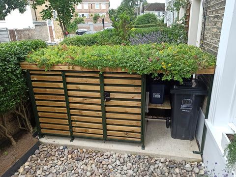 a wooden fence with plants growing on top and two trash cans in the middle next to it