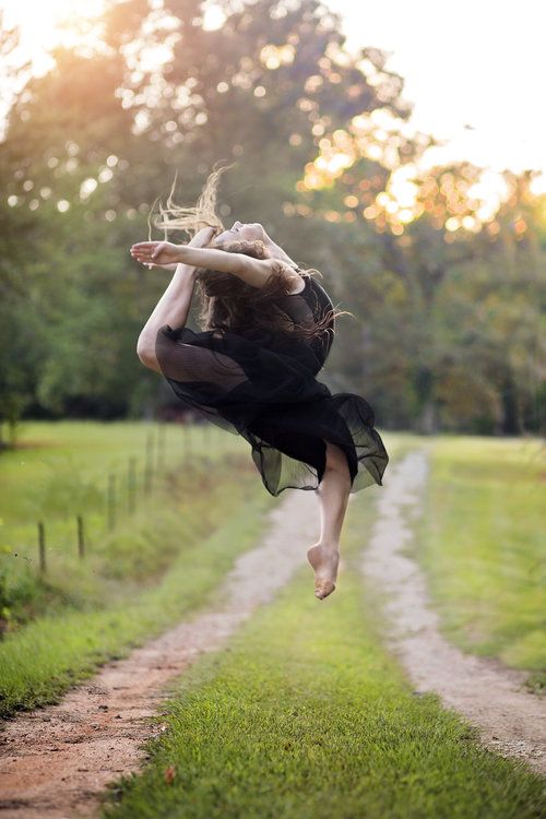 a woman is jumping in the air on a dirt road with grass and trees behind her