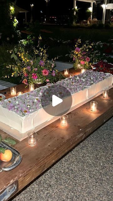 an outdoor buffet with candles and fruit on the table in front of flowers, bushes and trees