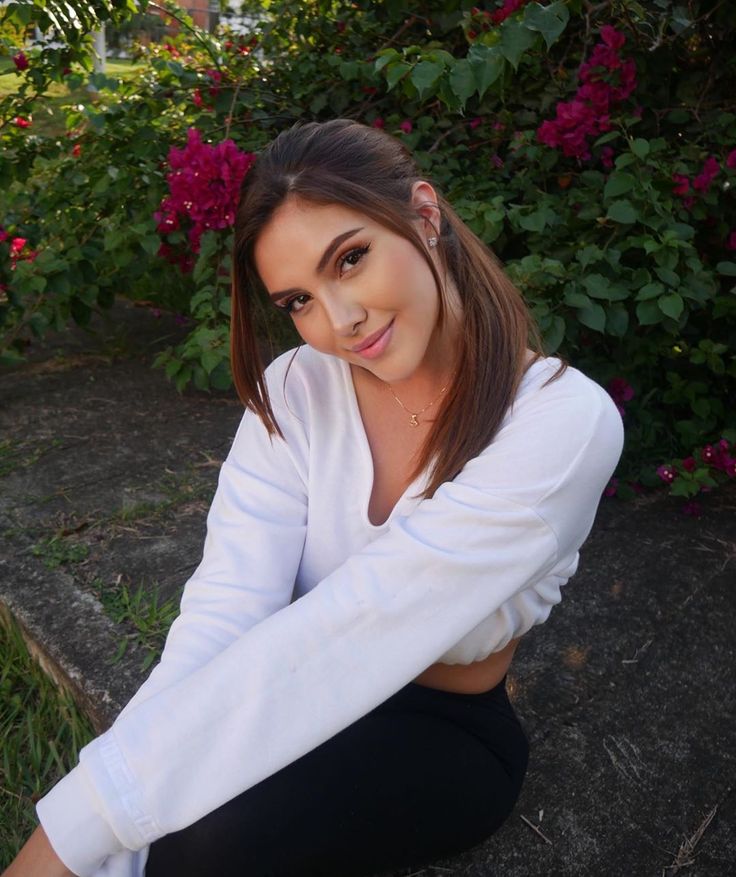 a beautiful young woman sitting on the ground in front of some bushes and pink flowers