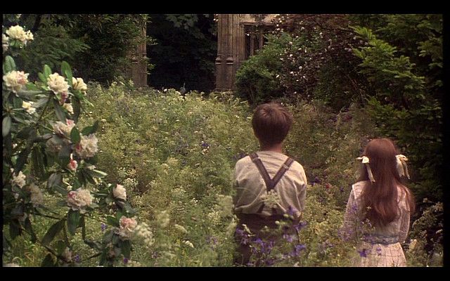 two people standing in the middle of a field with flowers and trees behind them, looking at an old building